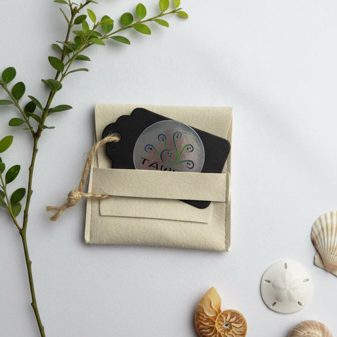 Pochette cadeau en tissu écru pour broche artisanale TAWUS, présentée avec feuillage et coquillages.