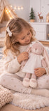 Une enfant assise sur un tapis tient la peluche lapin rose Léna de Bukowski Design, vêtue d’une robe blanche