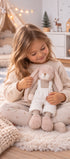 Une enfant assise sur un tapis tient la peluche lapin Lucian de Bukowski Design, vêtue d’une salopette blanche.