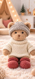  Peluche ours Robby de Bukowski Design assise sur un tapis, avec un pull écru, un bonnet gris et des chaussons rouges.