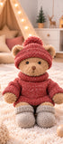  Peluche ours Steve de Bukowski Design assise sur un tapis, avec un pull rouge, un bonnet rouge à pompon et des chaussons gris