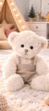 Peluche ours écru Wyatt de Bukowski Design assise sur un tapis dans une chambre, avec une salopette beige à bretelles