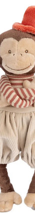 Peluche singe Winston de Bukowski Design sur fond blanc, les bras croisés, avec un pantalon beige, un haut rayé et un chapeau rouge