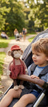 Un enfant en poussette tient la peluche singe Winston de Bukowski Design, en salopette rouge et chapeau assorti, dans un parc.