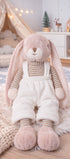 Peluche lapin Lucian de Bukowski Design, en salopette blanche et haut rayé, assise sur un tapis dans une chambre d’enfant.